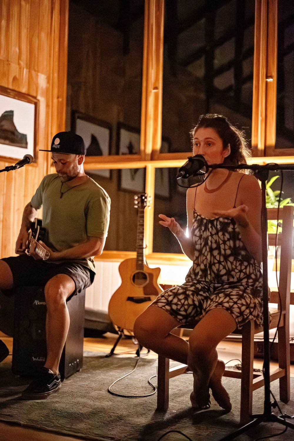 Acoustic musicians perform on stage in a warmly lit wooden room with a guitar visible in the background.