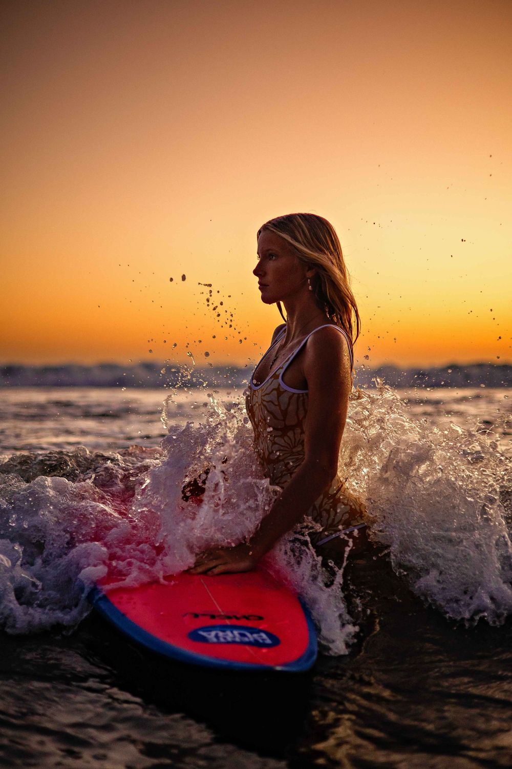 Surfer rides waves on a red bodyboard against vibrant orange sunset sky with splashing water silhouettes.