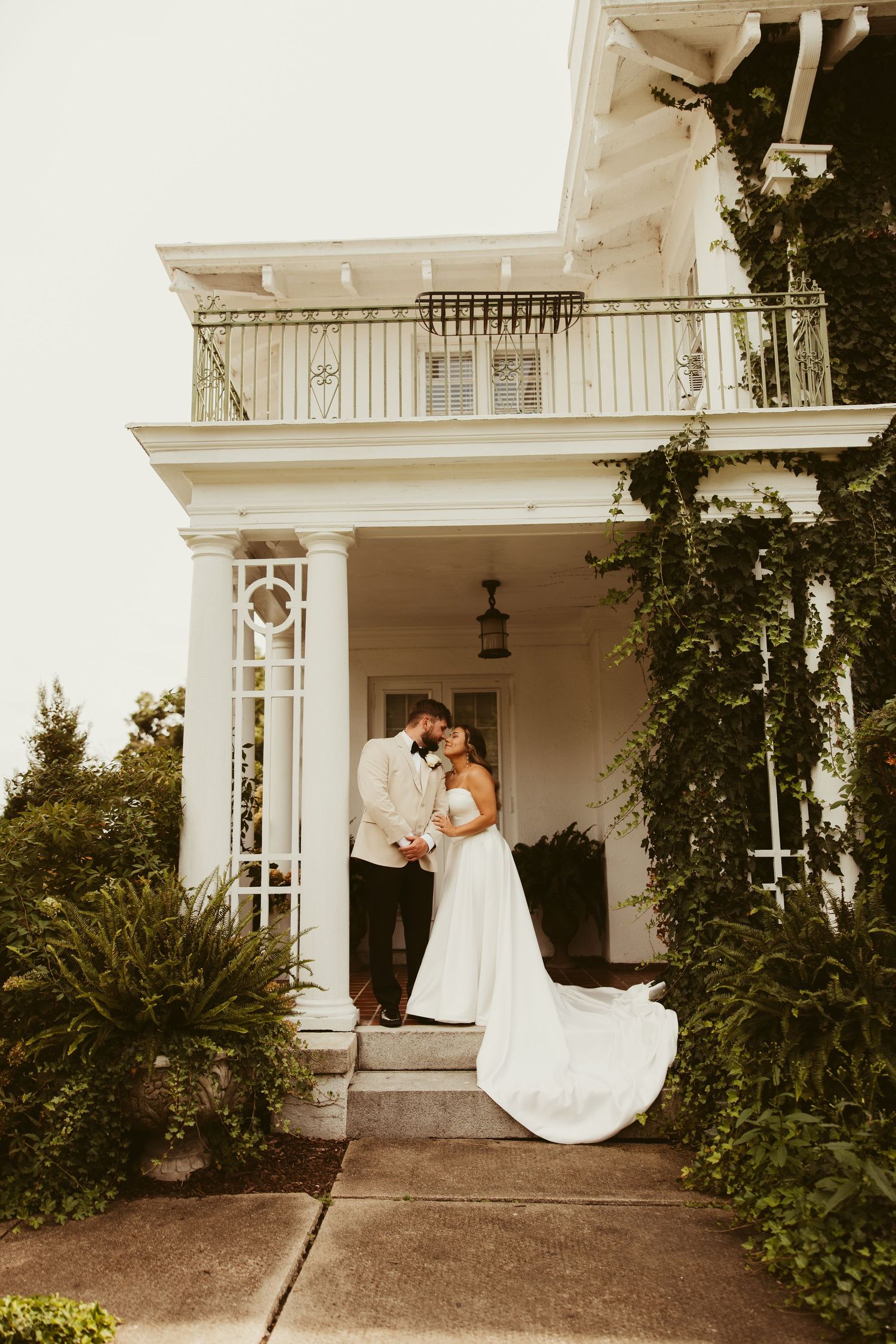 Bride and groom share romantic moment on steps of elegant white Southern mansion with ivy-covered columns.