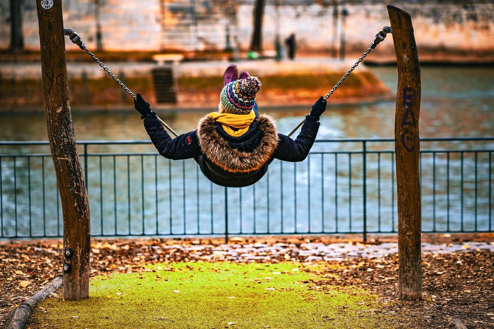Someone wearing winter clothing swings joyfully between two trees by a waterfront on an autumn day with fallen leaves.