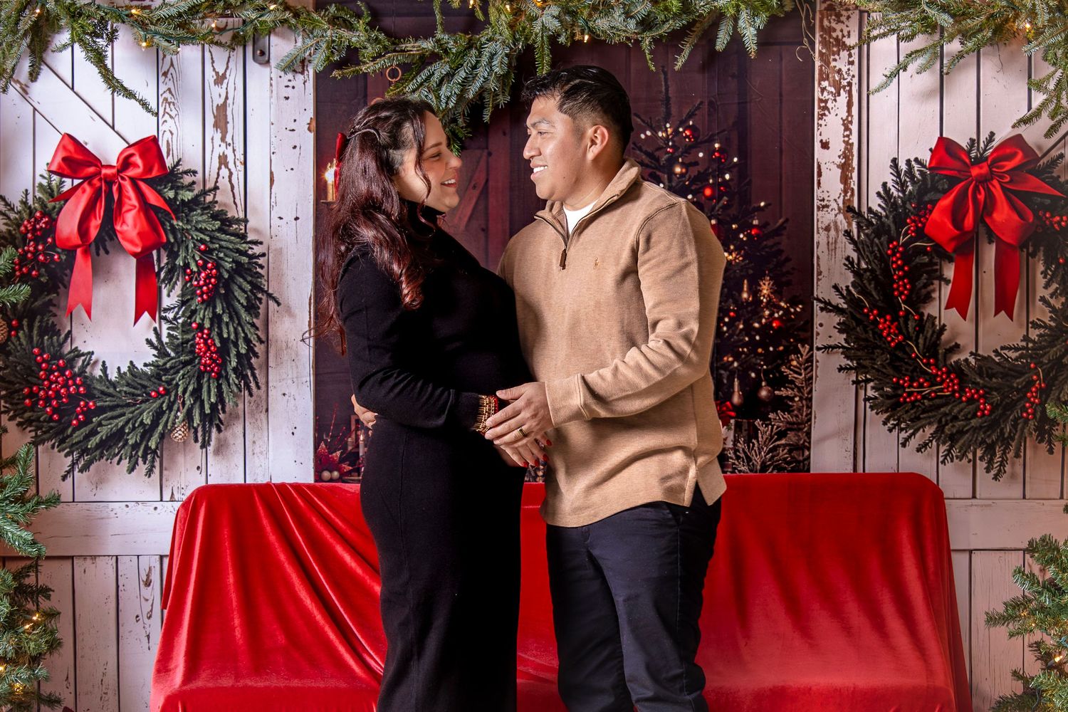 Romantic couple embraces in front of white wall decorated with festive Christmas wreaths and red bows.