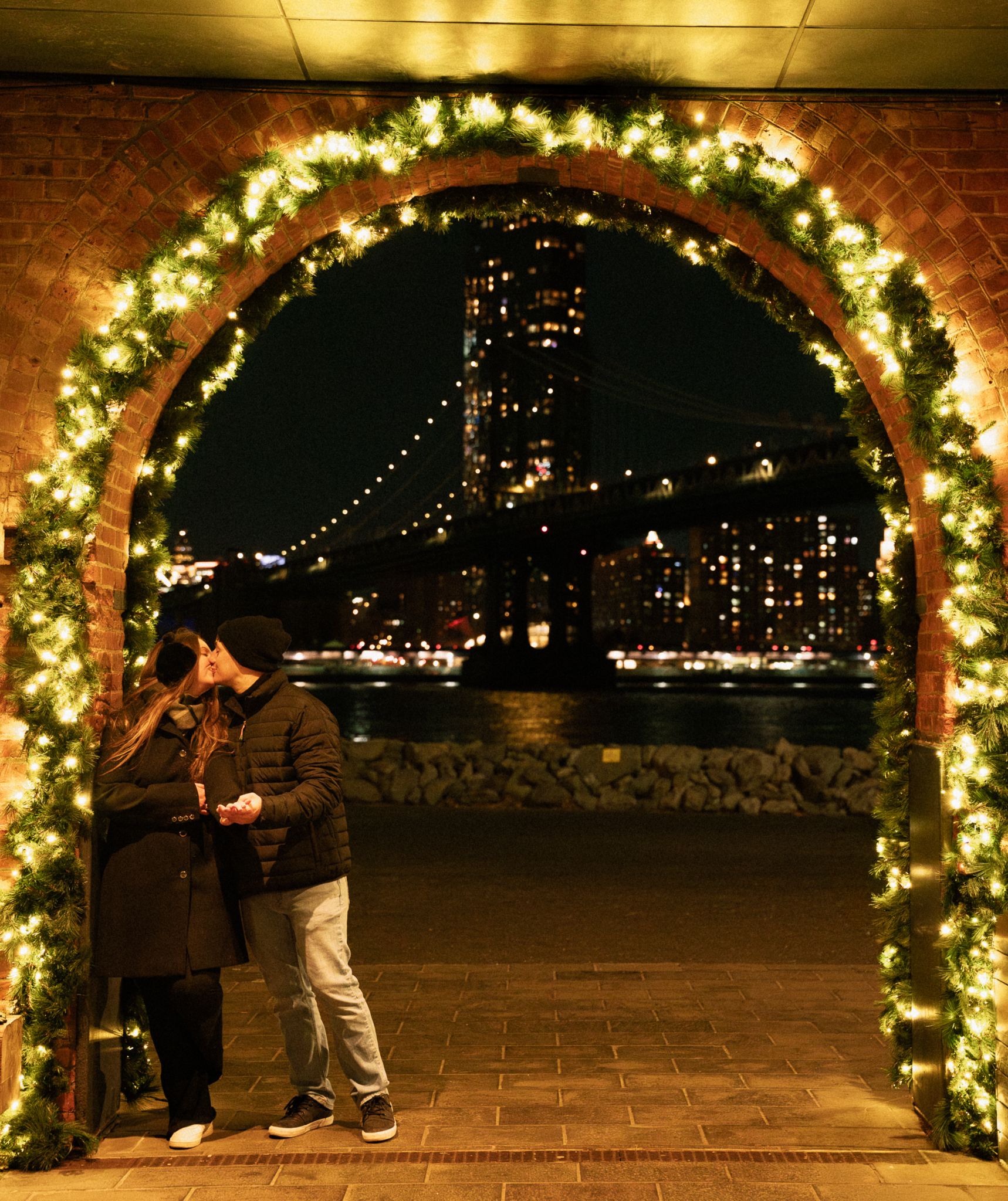 A romantic moment framed by an archway adorned with holiday garland overlooking the city lights.