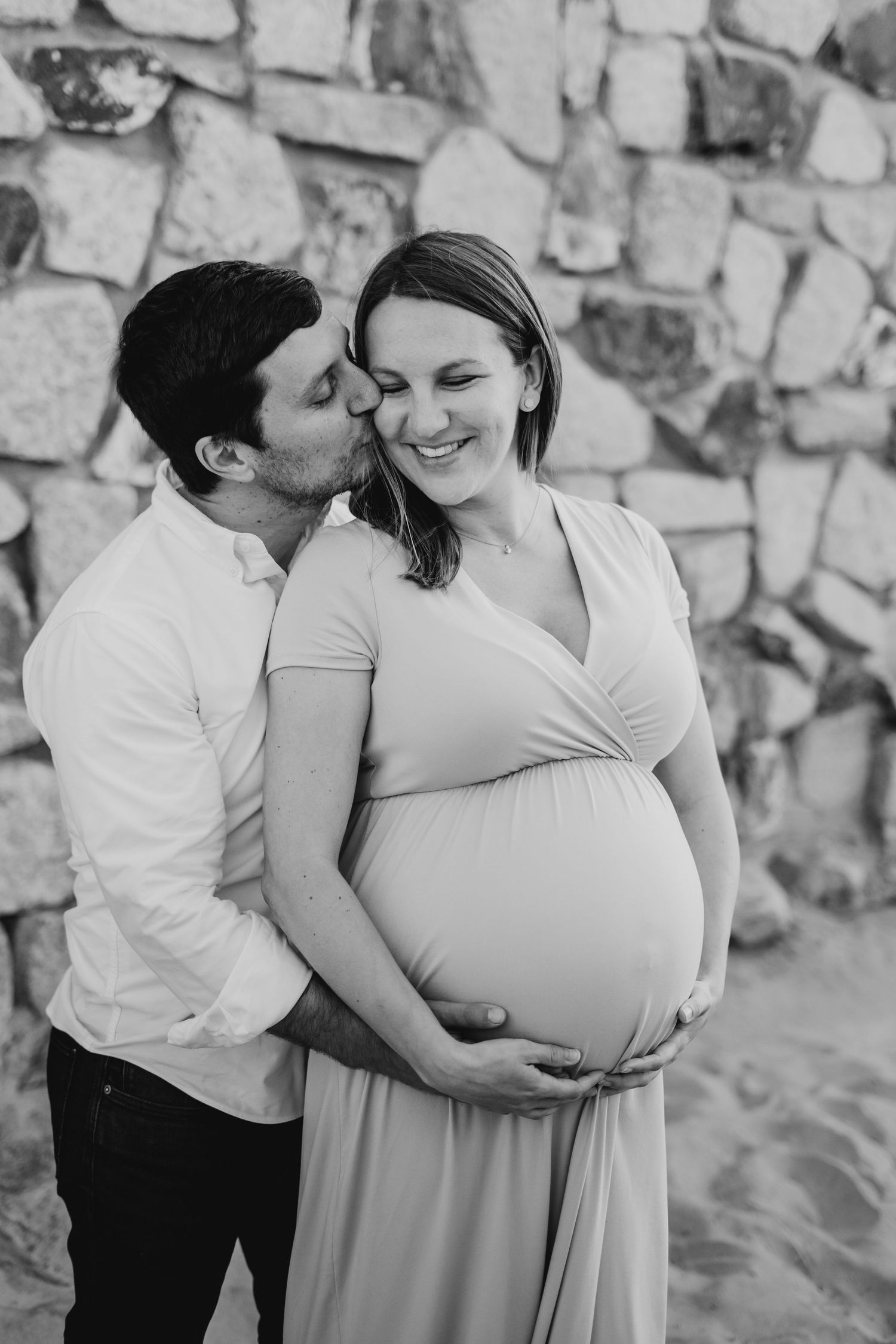 Black and white maternity photo of couple at Coronado Beach with rock wall in background