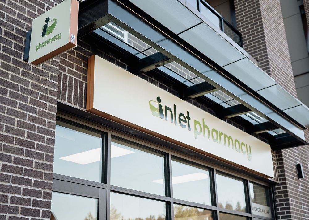 A modern pharmacy storefront with glass windows and a green logo sign mounted on brick exterior walls.