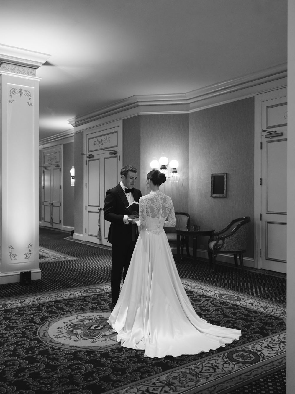 Elegant wedding couple in formal attire photographed in a hotel hallway with ornate carpeting and wall sconces.