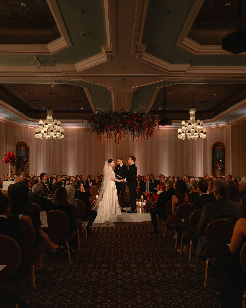Wide view of a wedding ceremony in a grand ballroom with chandeliers and rows of seated guests.