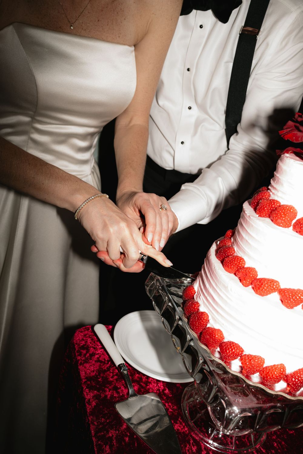 Close-up shots of hands cutting into a white wedding cake decorated with red strawberries.