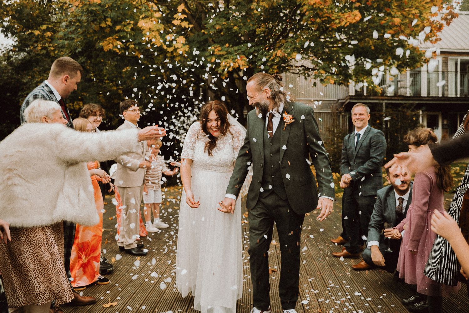 Wedding guests shower newlyweds with white flower petals during an autumn outdoor celebration.