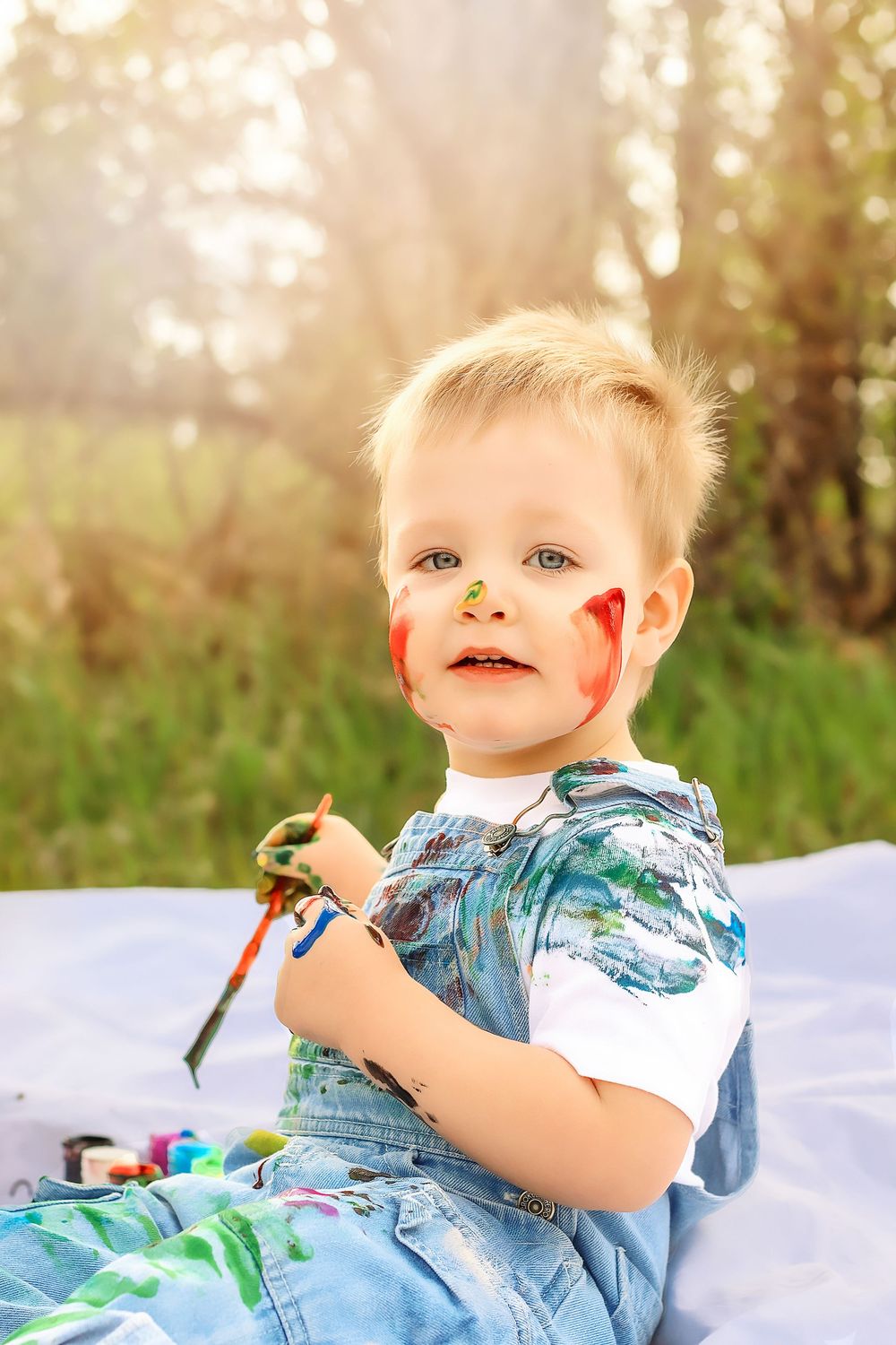 A young child lies on a white blanket outdoors in a floral shirt with sunlit trees in the background in Williston, North Dakota.