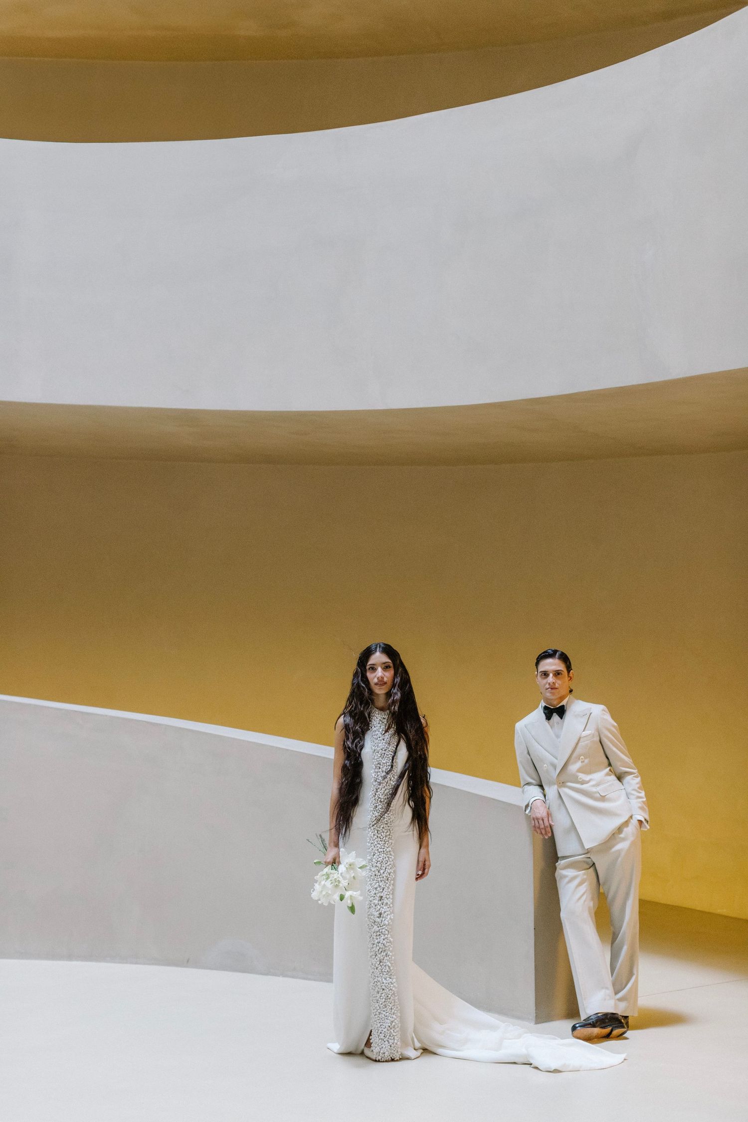 Wedding couple in white attire posing dramatically on a curved white and yellow backdrop.