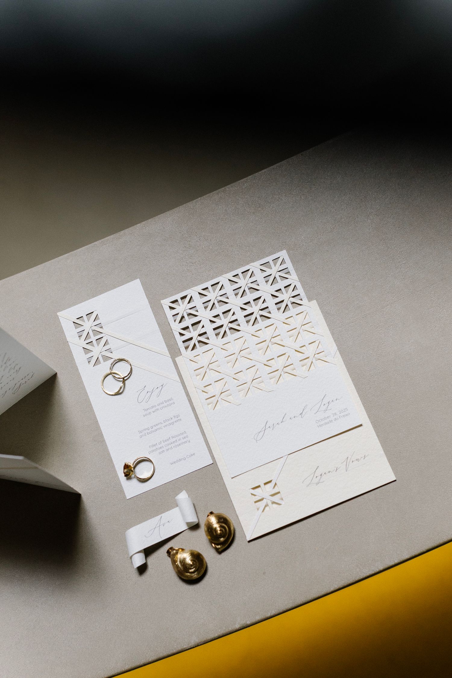 Minimalist white wedding invitation suite with geometric cutout pattern and gold wedding rings on gray surface.
