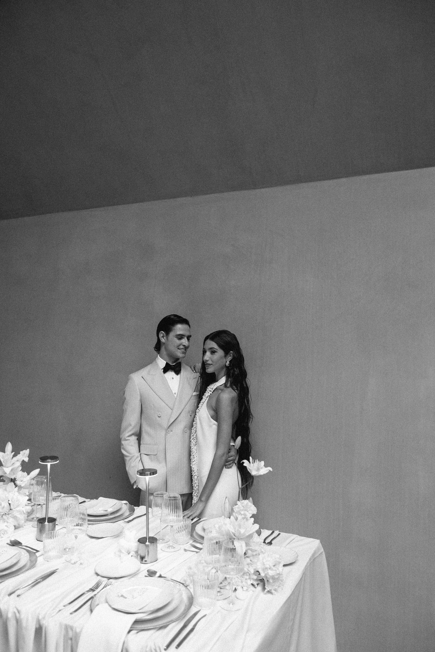 A black and white wedding photo of a couple standing by an elegantly set table with white linens and floral arrangements.