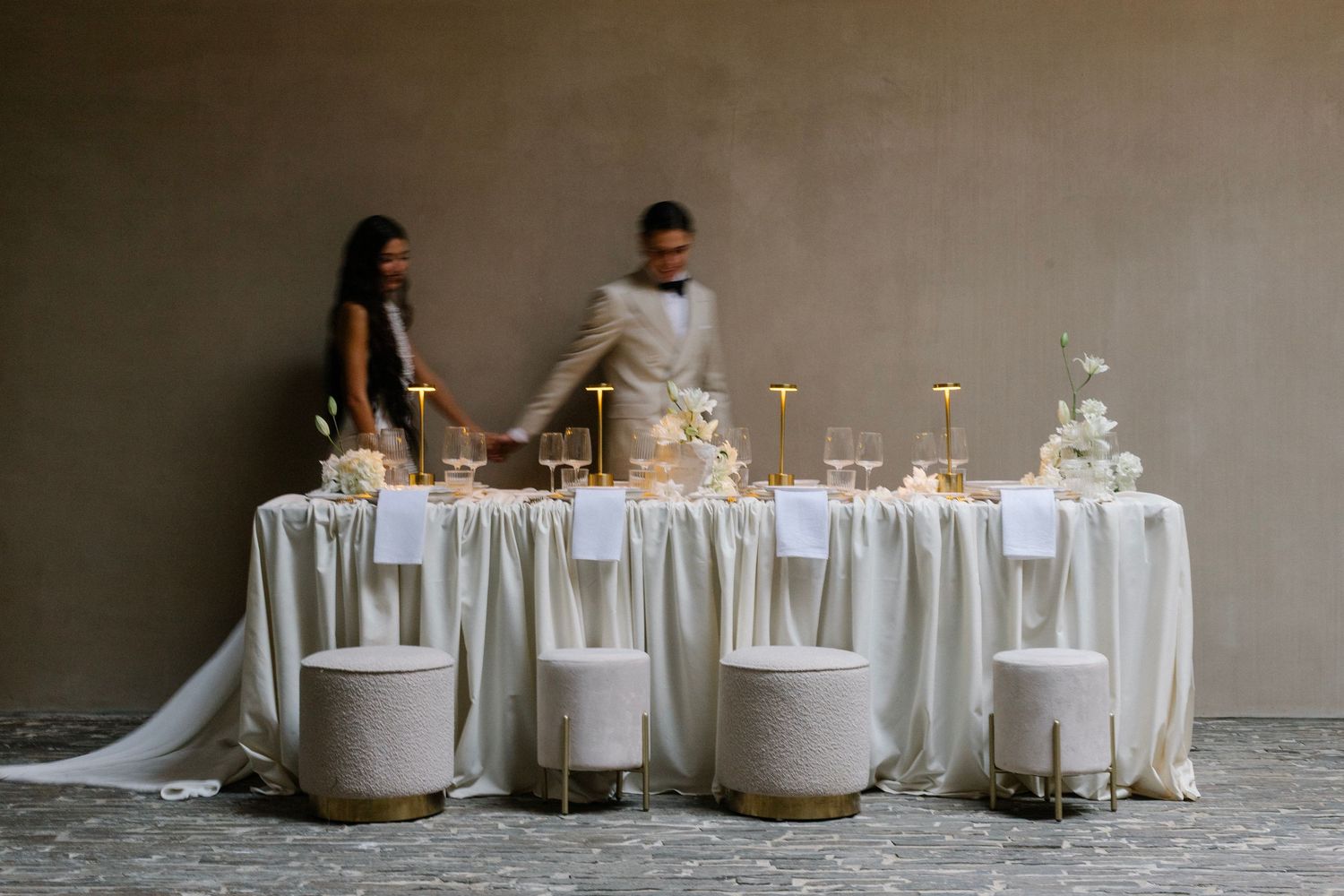 Elegant wedding reception table setup with white linens and glassware being arranged by two people.
