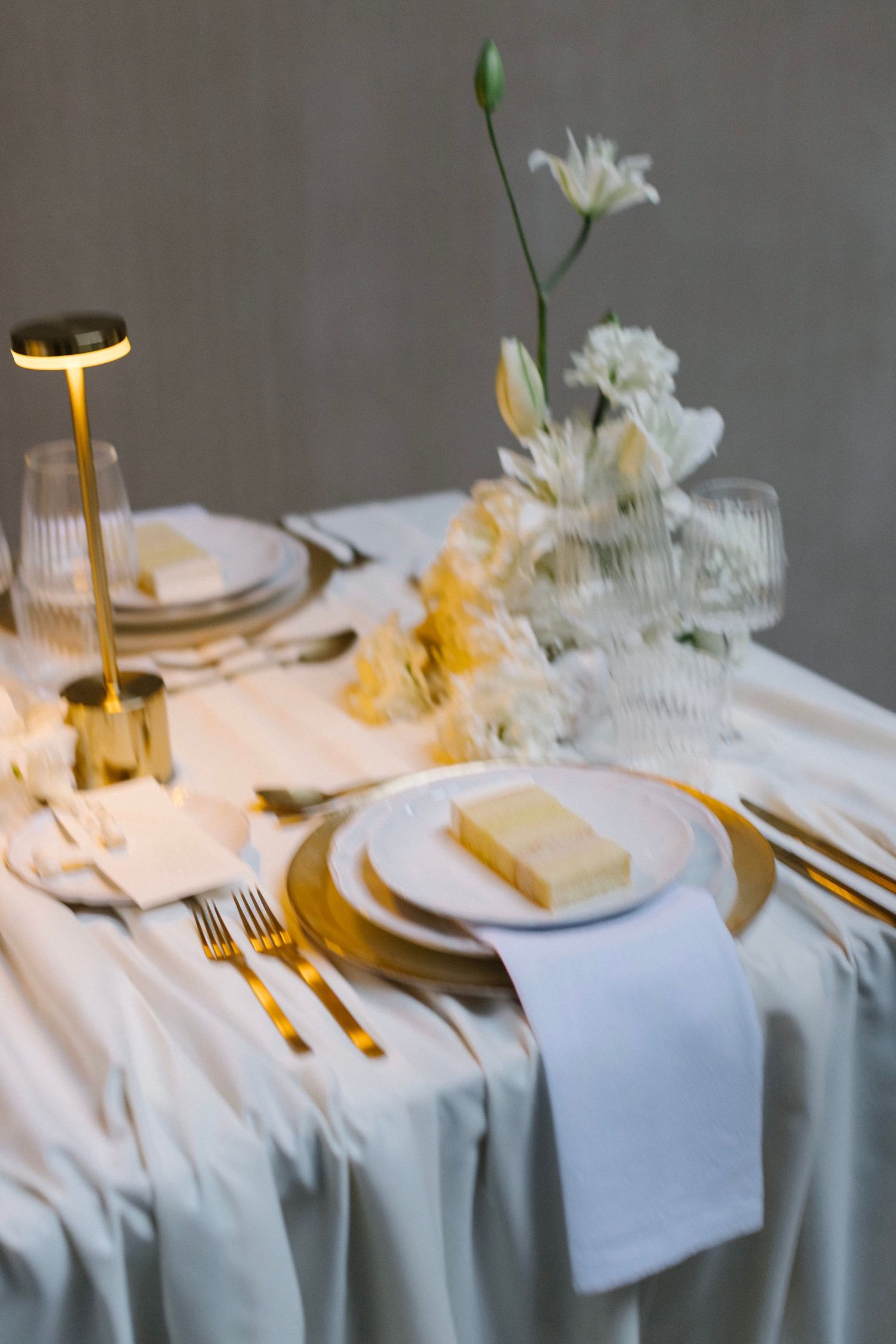 Elegant table setting with gold cutlery, white plates, napkins and delicate floral centerpiece in crystal vases on white tablecloth.