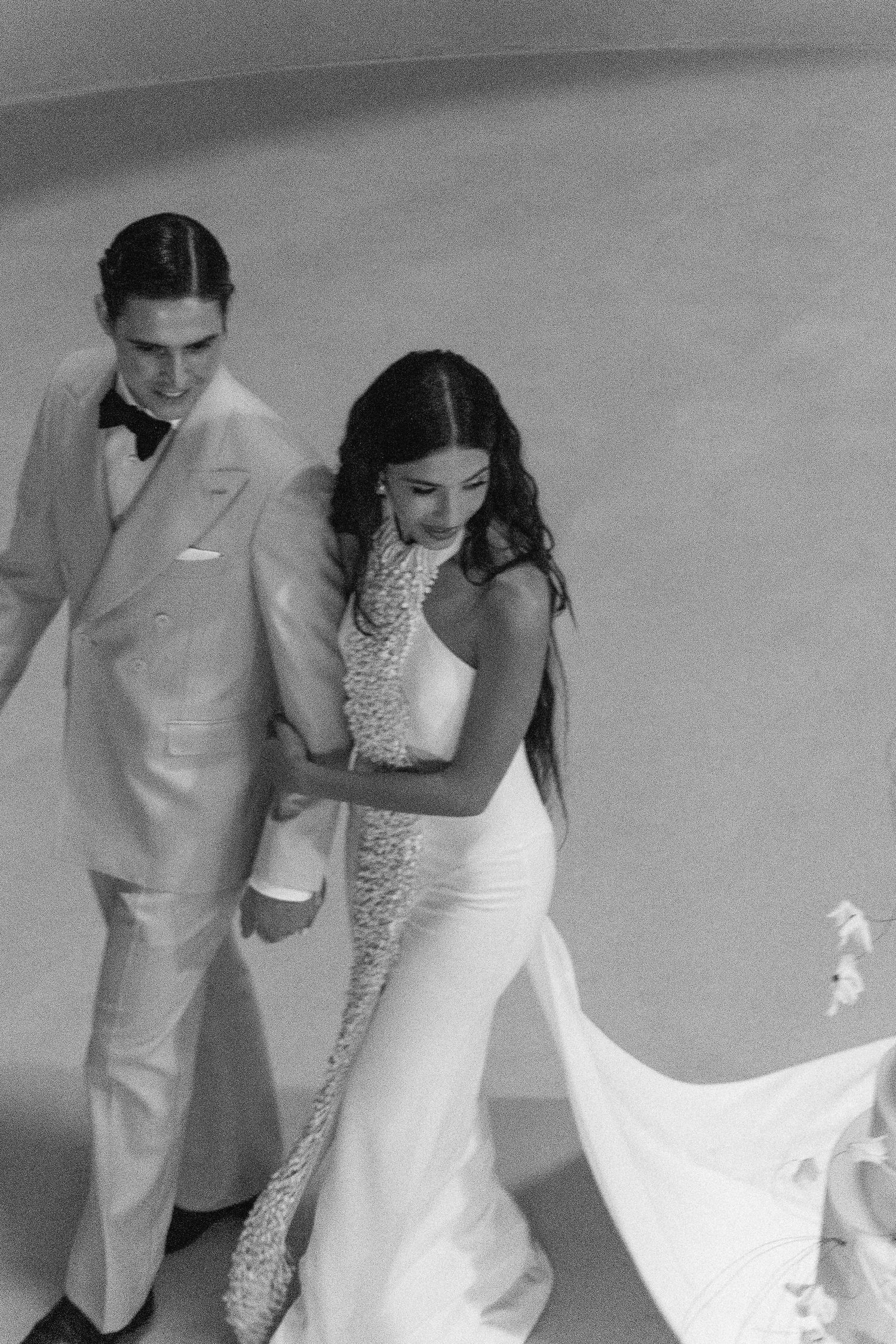 Black and white photo of a couple in formal attire walking together with a flowing white dress train.