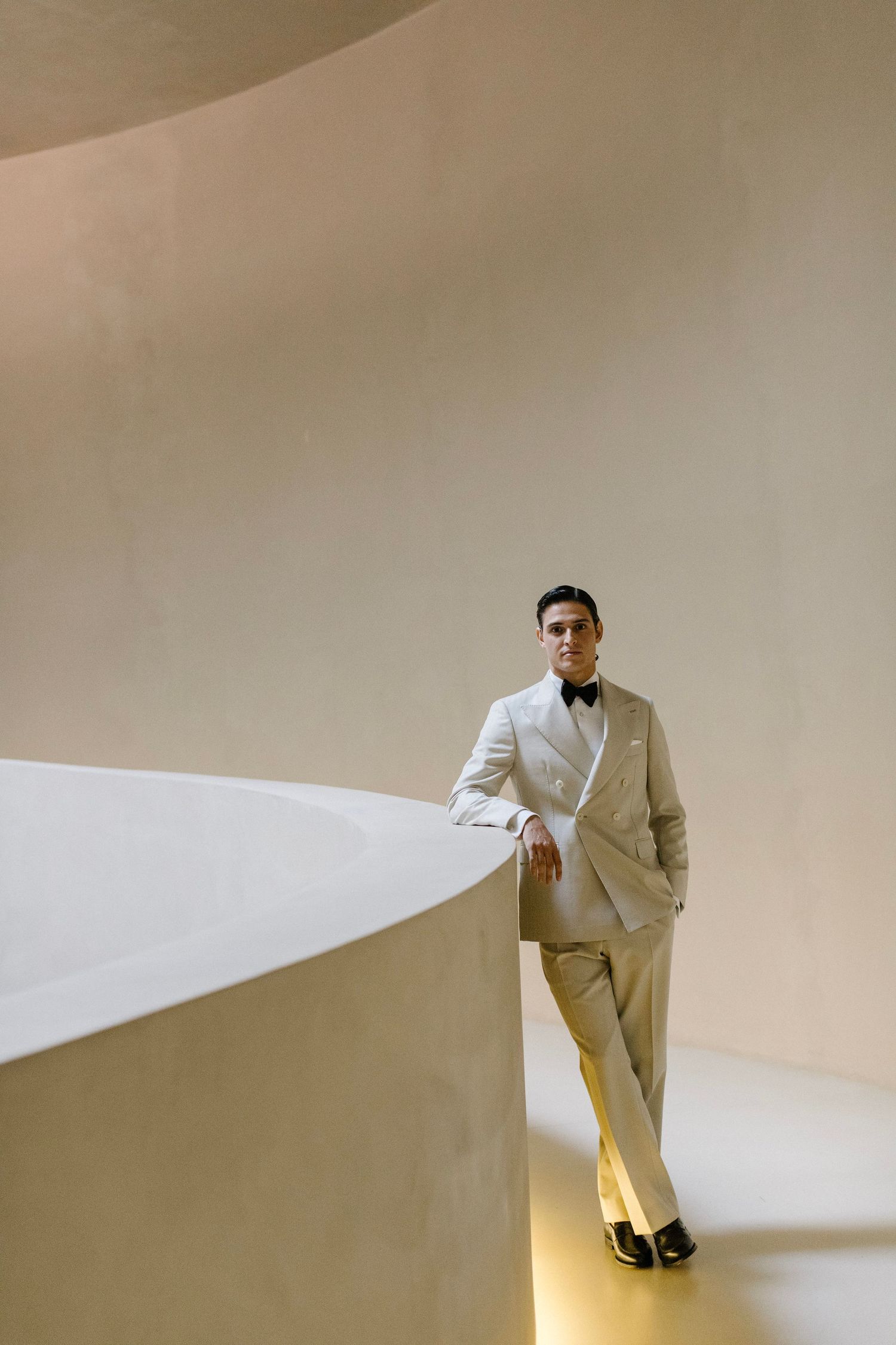 Elegant figure in cream-colored tuxedo and bow tie leans against curved white wall in minimalist architectural setting.