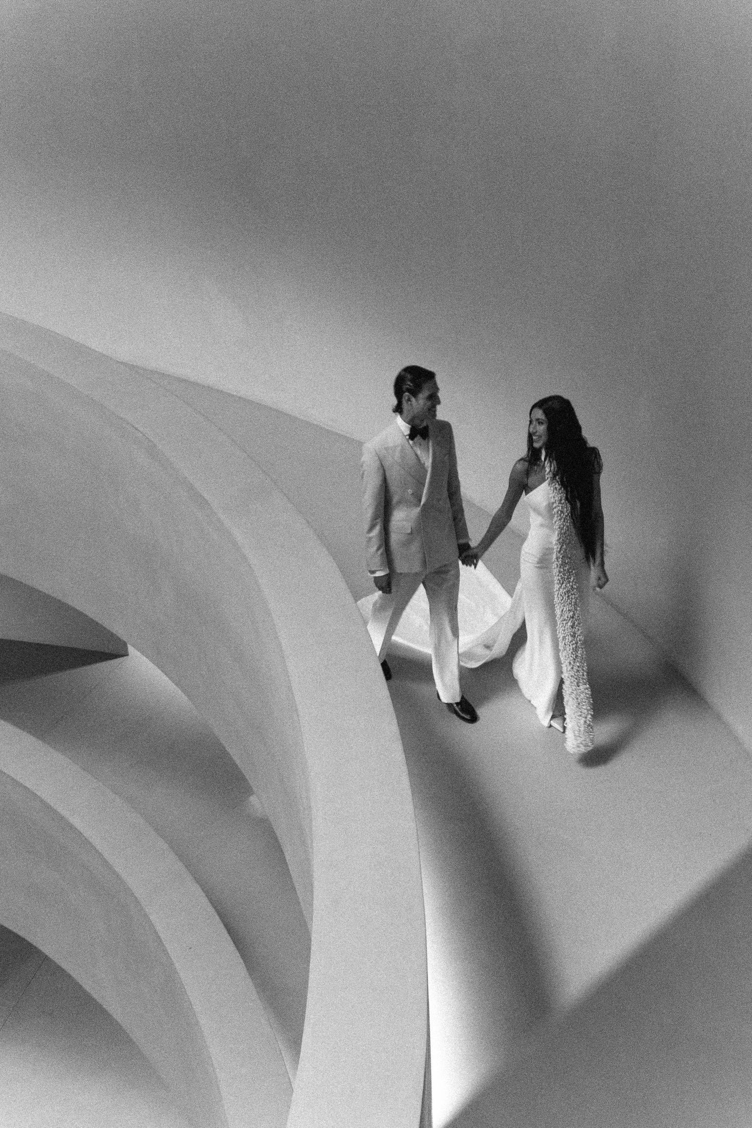 A black and white artistic wedding photo of a couple walking hand in hand along a curved white architectural structure.