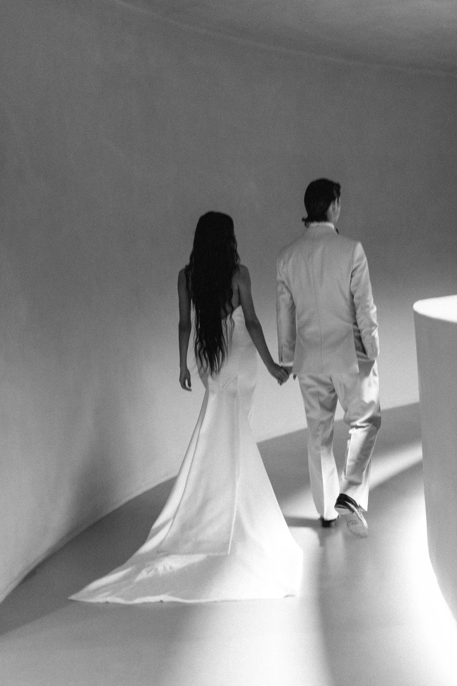 A couple in wedding attire holding hands while walking up curved white stairs in dramatic black and white lighting.