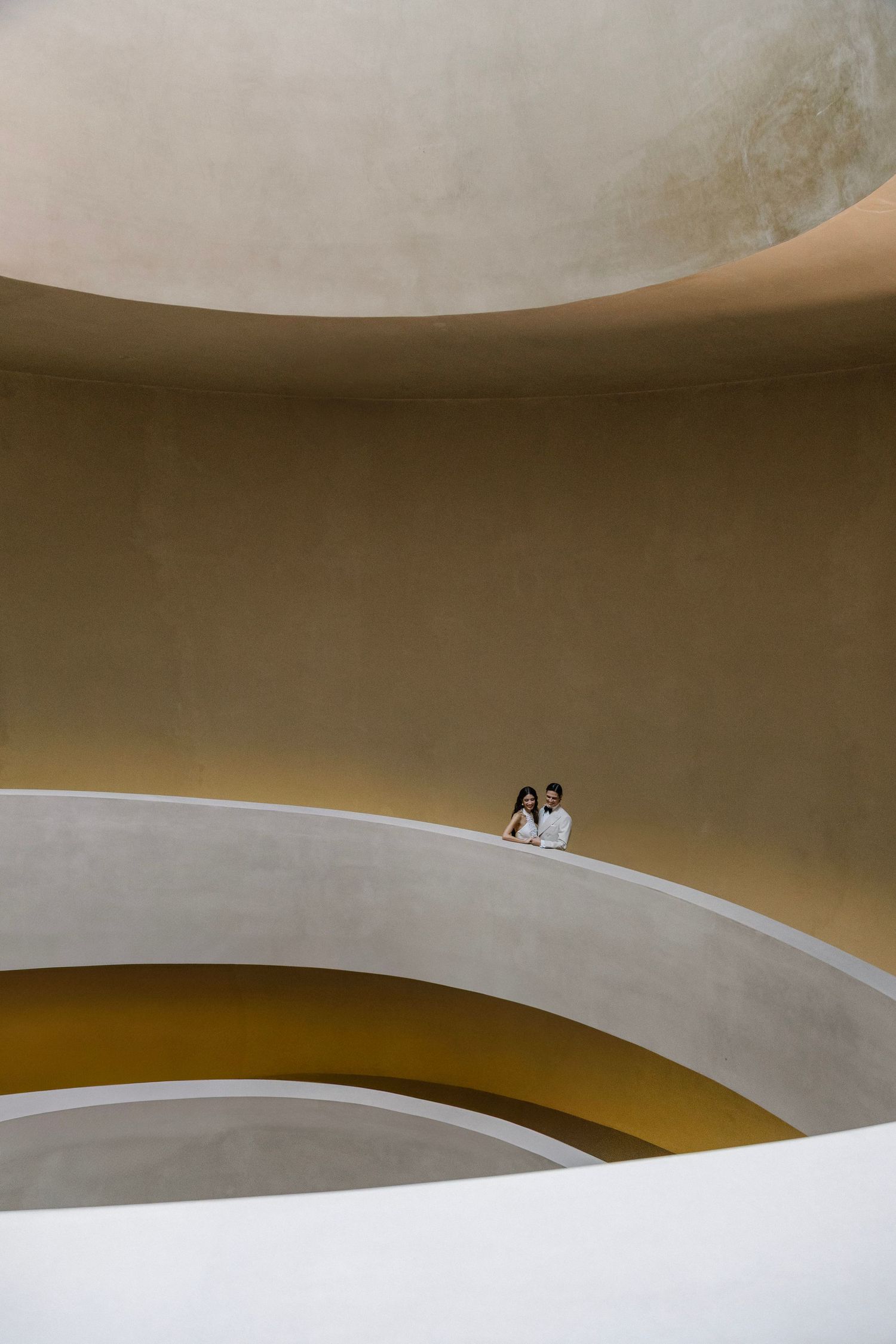 Two figures stand on a curved white architectural ramp against warm beige walls in a modern minimalist space.
