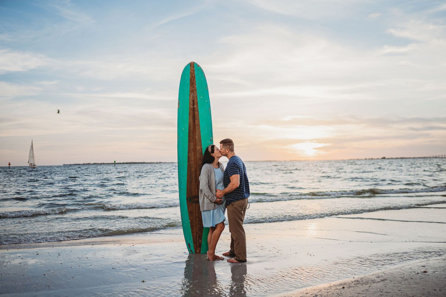 A couple with a surfboard shares a romantic moment on a beach at sunset against a serene ocean backdrop.