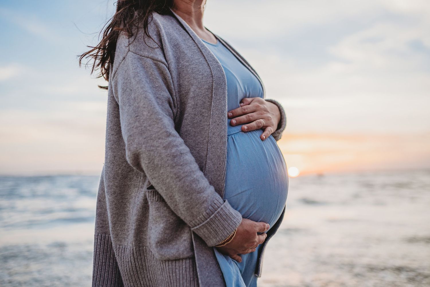 A pregnant woman in a blue dress and gray cardigan stands at the beach during sunset, cradling her baby bump.