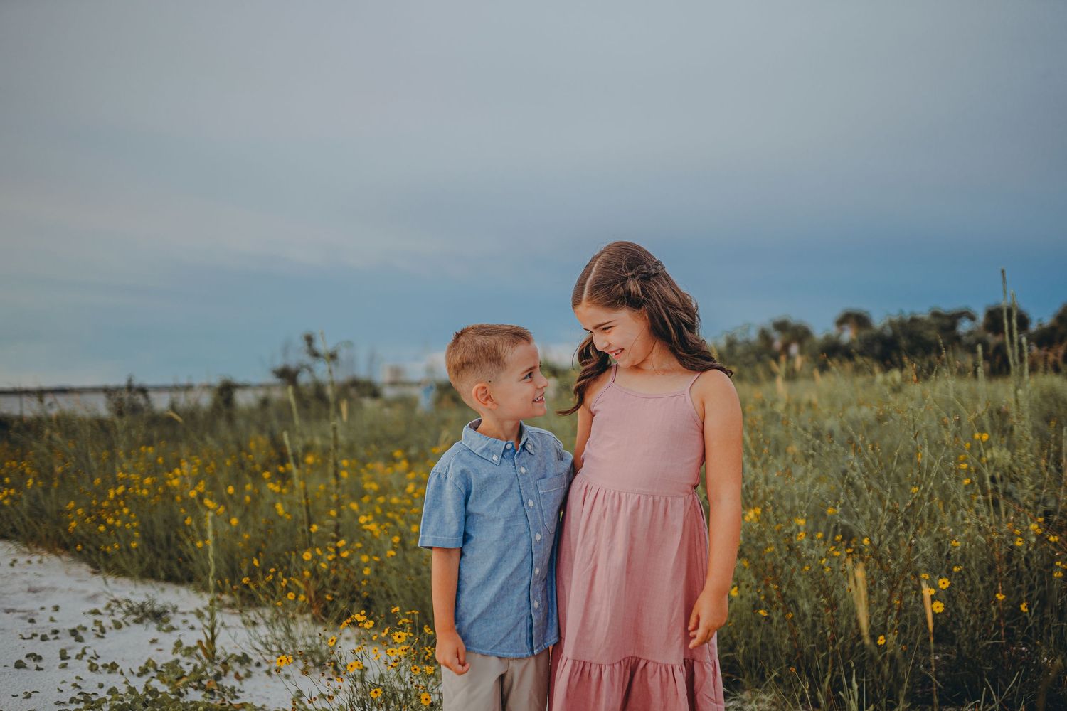 Two children stand together in a field of yellow wildflowers at sunset wearing a blue shirt and pink dress.