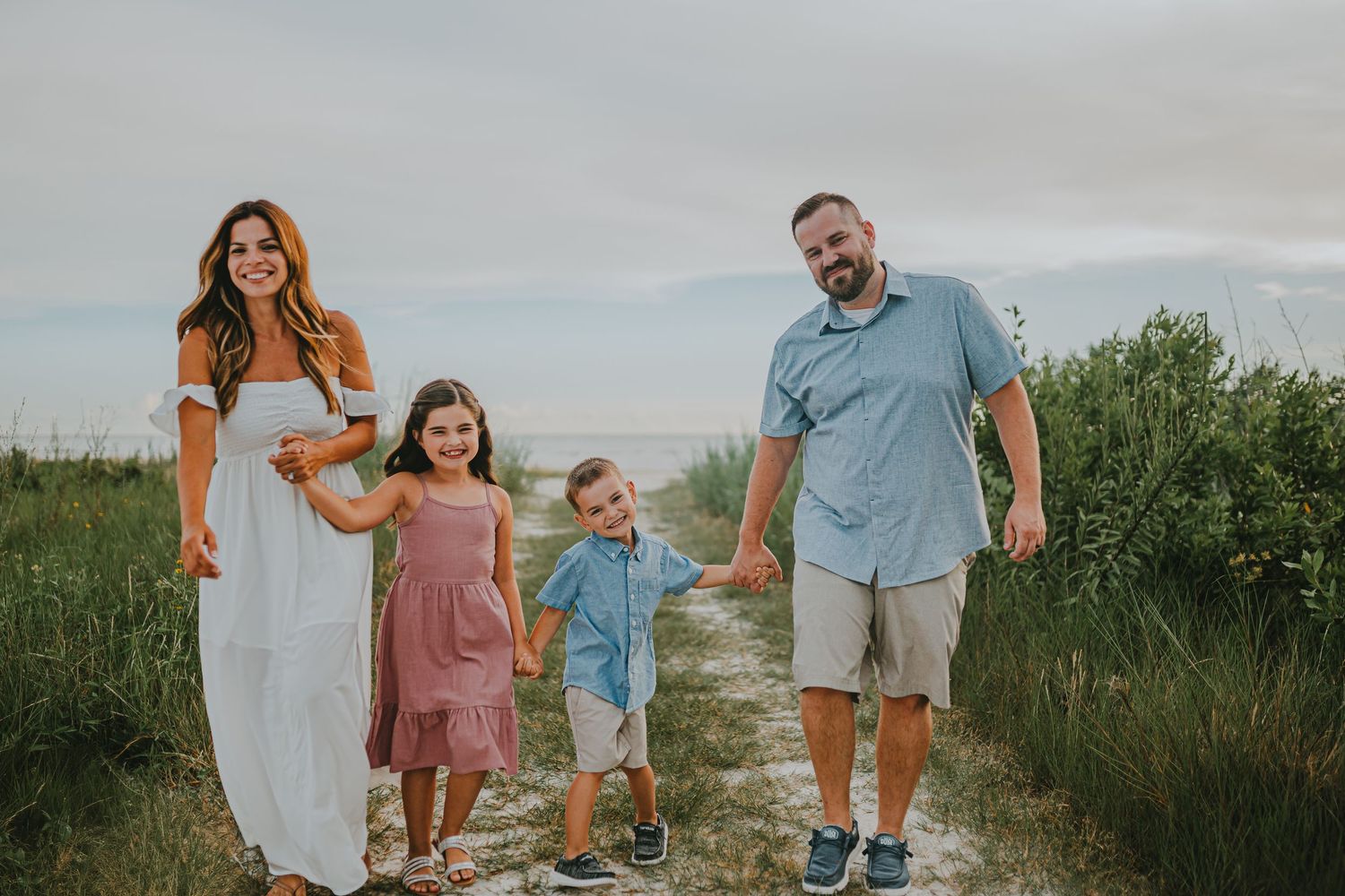 A family walks together on a sandy beach path through green coastal vegetation during a sunny day.