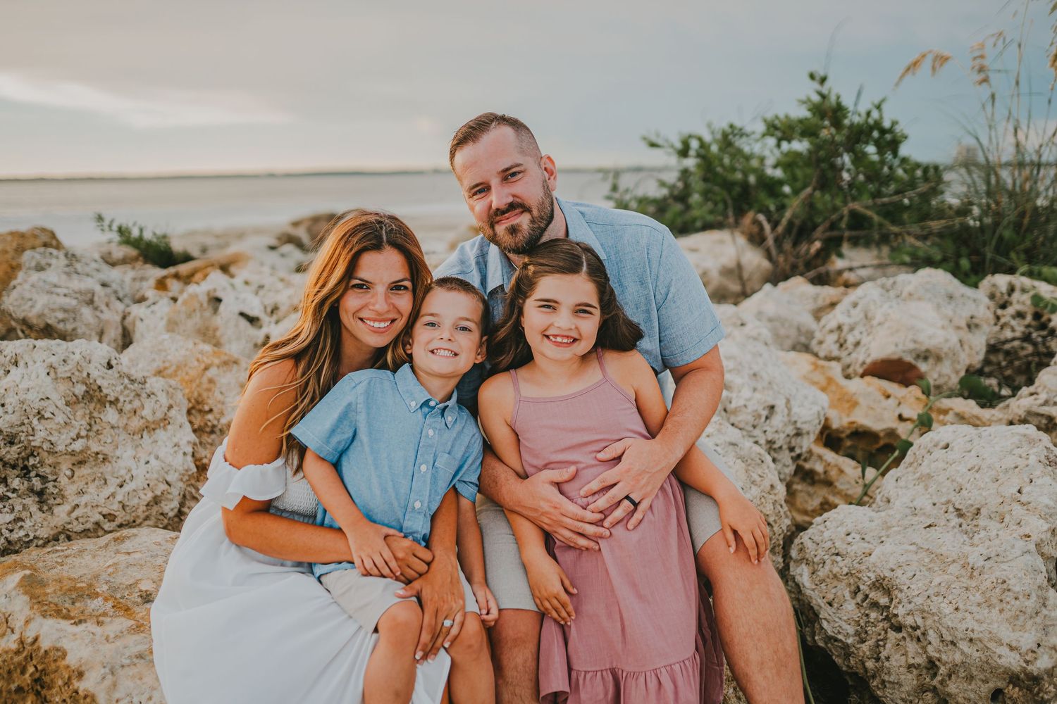 Family poses together on rocky beach terrain wearing coordinated blue and pink outfits during sunset photo session.