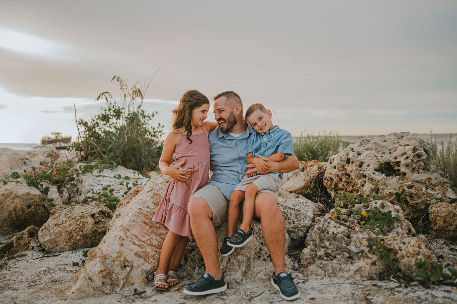 Family wearing coordinating coral and blue outfits sits together on rocky beach during sunset photoshoot.