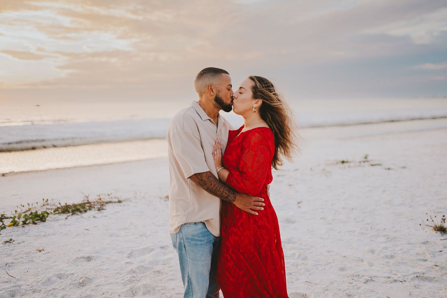 A couple in a red dress and gray suit embrace on a pristine white sandy beach during a romantic photoshoot.