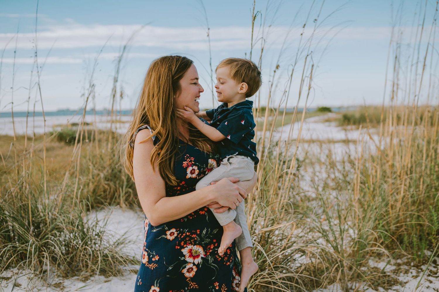 A couple embraces in a serene coastal setting with tall grass swaying in the breeze at sunset.