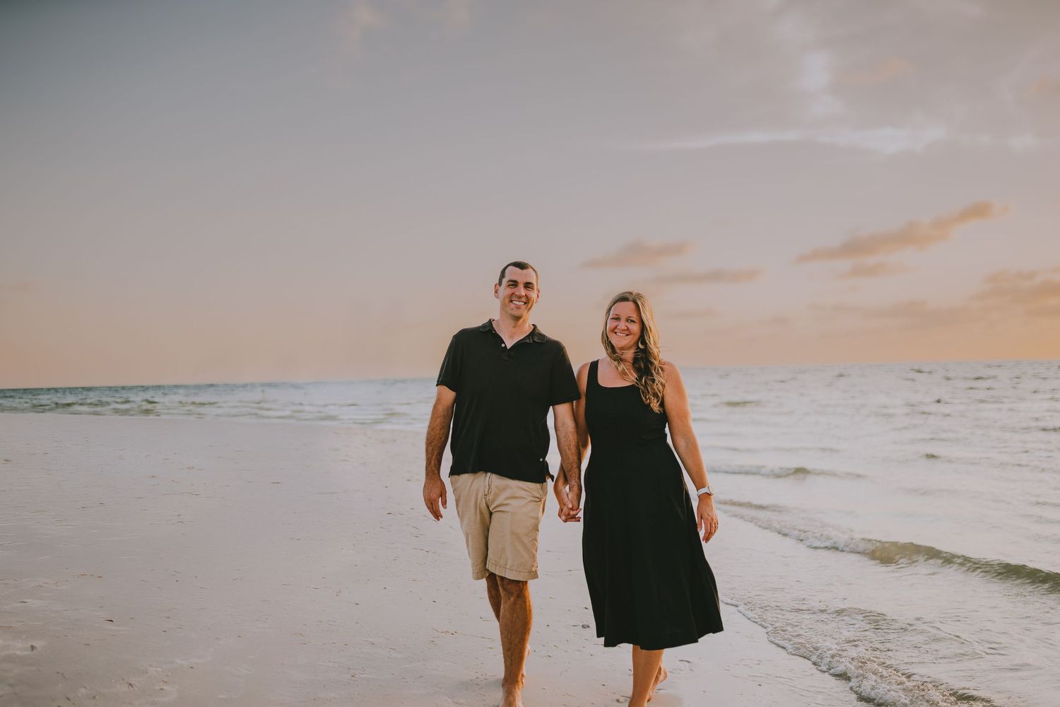 A couple walking together on a white sandy beach at sunset with ocean waves in the background.