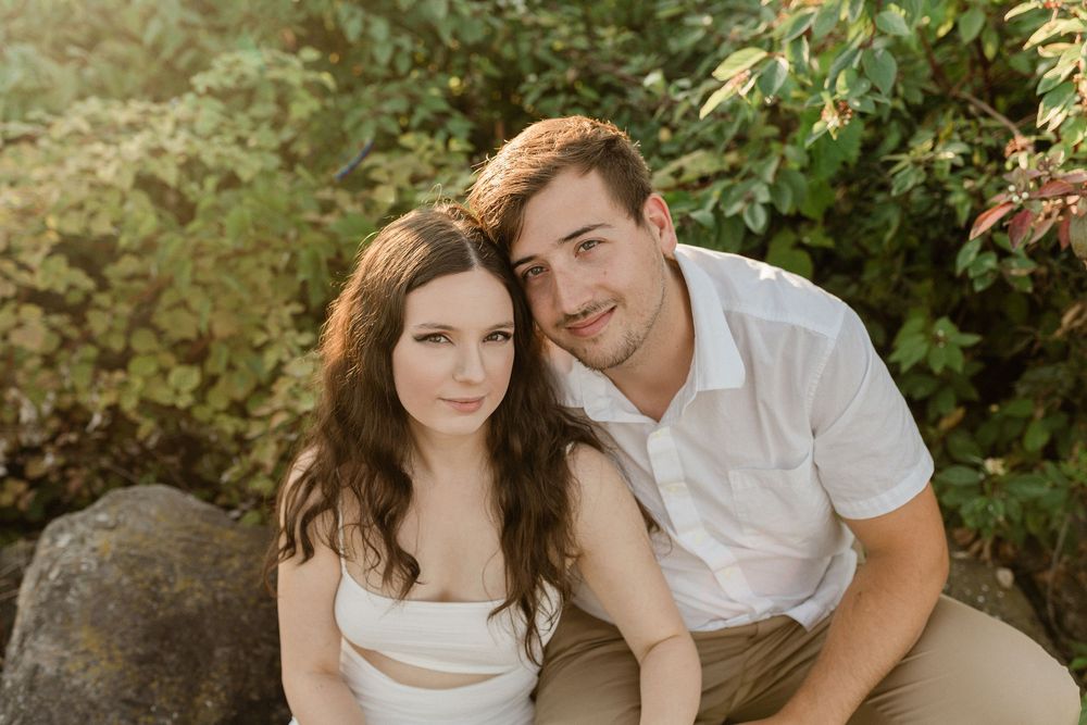 Couple in light colored outfits share tender moments among lush green foliage outdoors.
