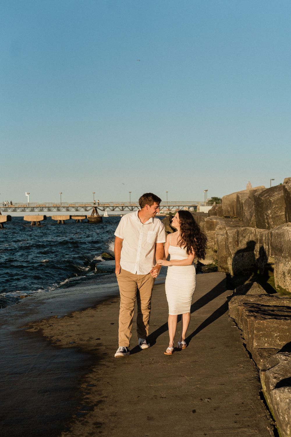 A couple in white and khaki outfits walking along a stone pier by the ocean at sunset.