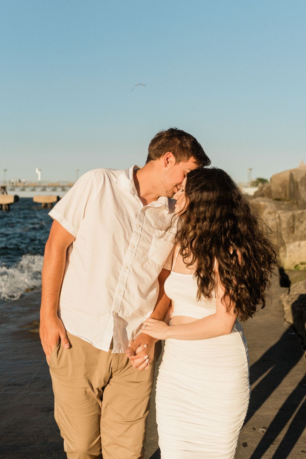Couple embracing on a coastal walkway with ocean and buildings visible in the background.