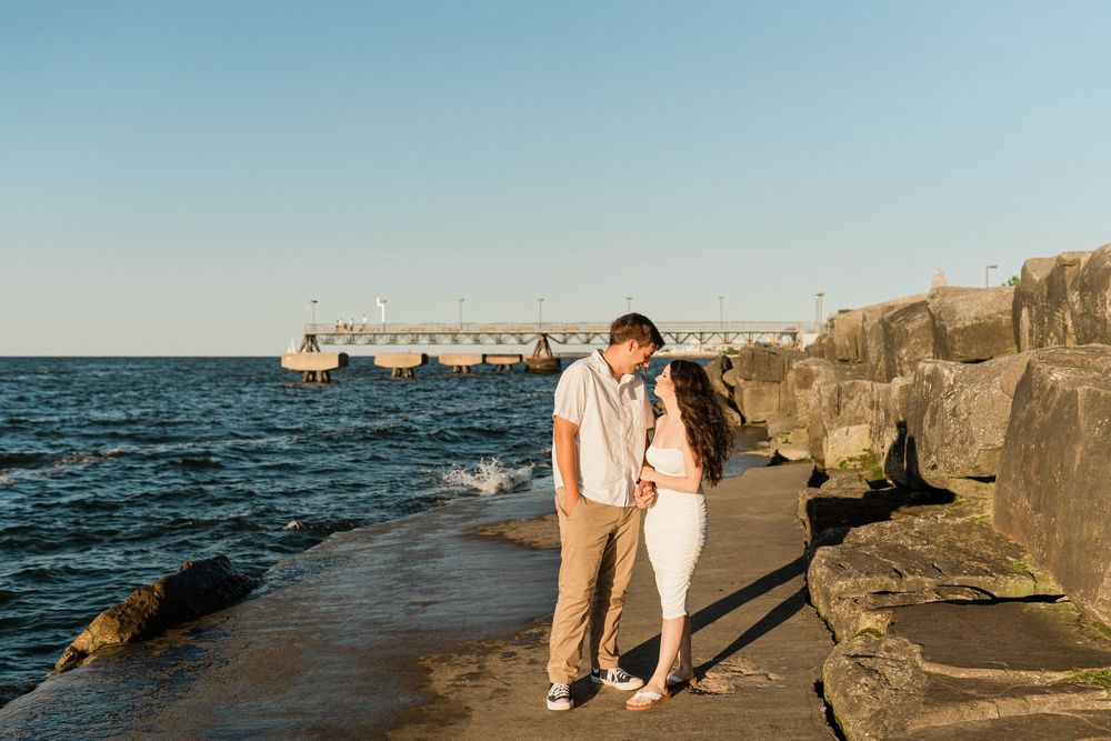 Couple walking along rocky shoreline with pier in background.