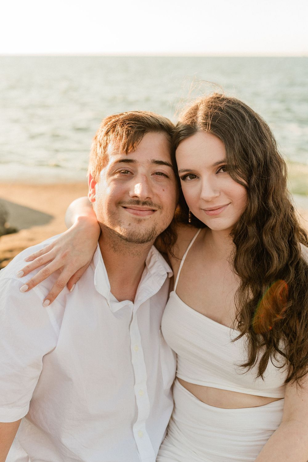 A romantic couple in white outfits shares intimate moments during a beach photoshoot at sunset.