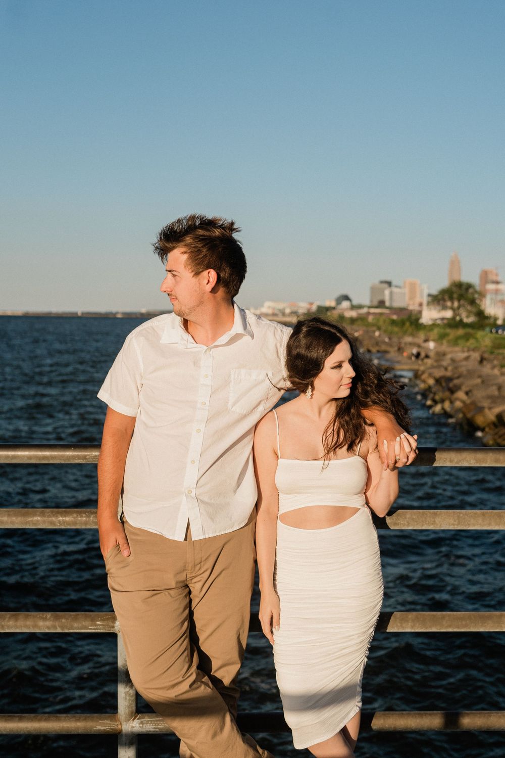 Romantic couple in white and khaki outfits share intimate moments along a waterfront boardwalk at sunset.