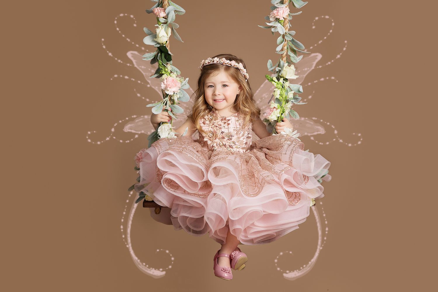 A child in a pink tulle dress sits on a floral swing against a brown backdrop in a dreamy portrait setting.