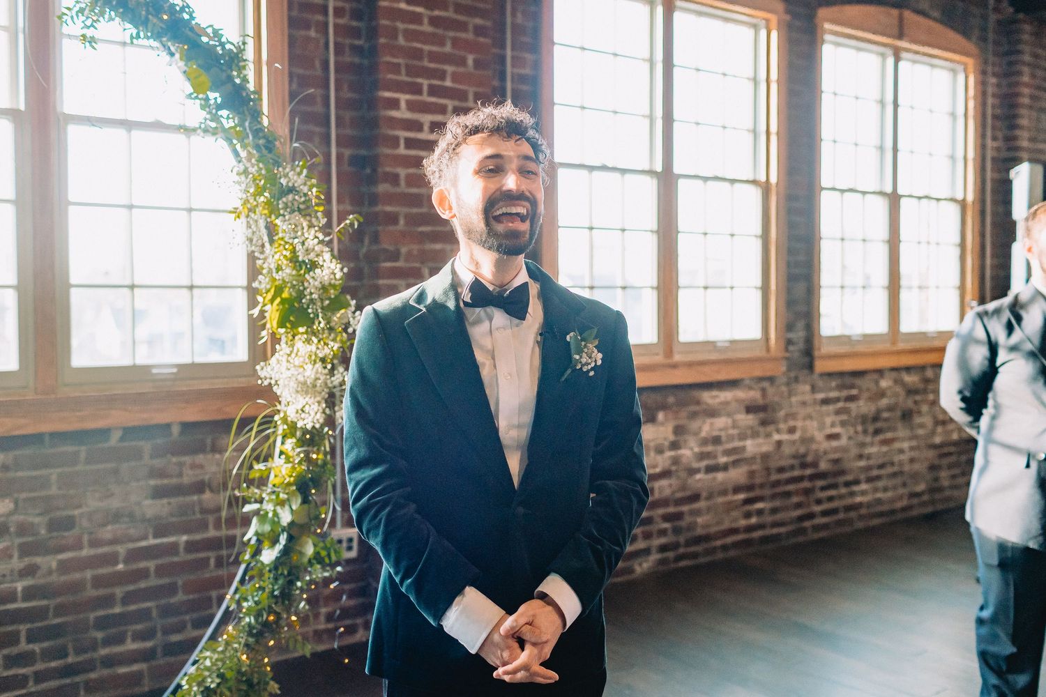 A person in a black tuxedo and bowtie stands smiling in front of brick walls and large windows at an indoor venue.