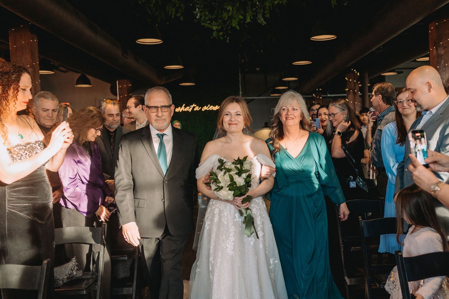 Wedding ceremony guests line both sides of an indoor aisle as a couple makes their entrance at an evening celebration.