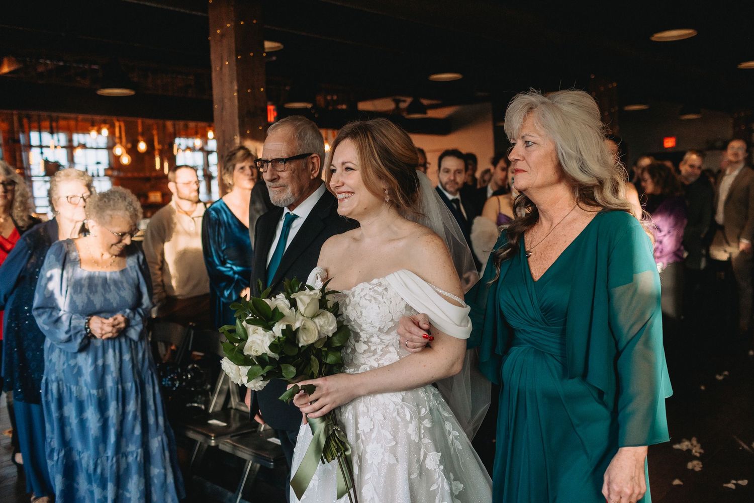 Wedding party members in teal dresses dance and celebrate with bride in white gown at indoor reception venue.