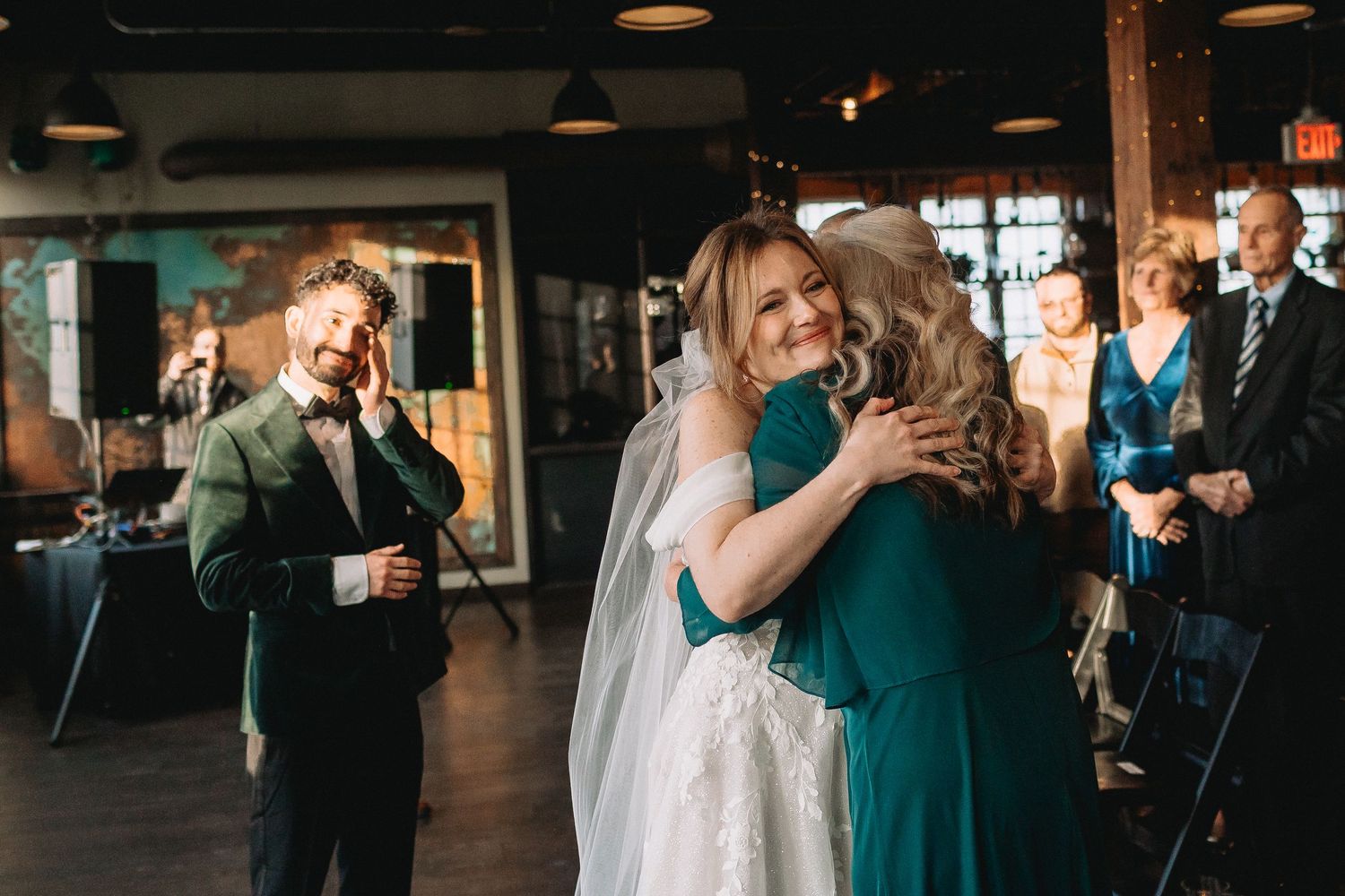 A couple embraces on the dance floor during a wedding reception with guests in teal dresses in the background.