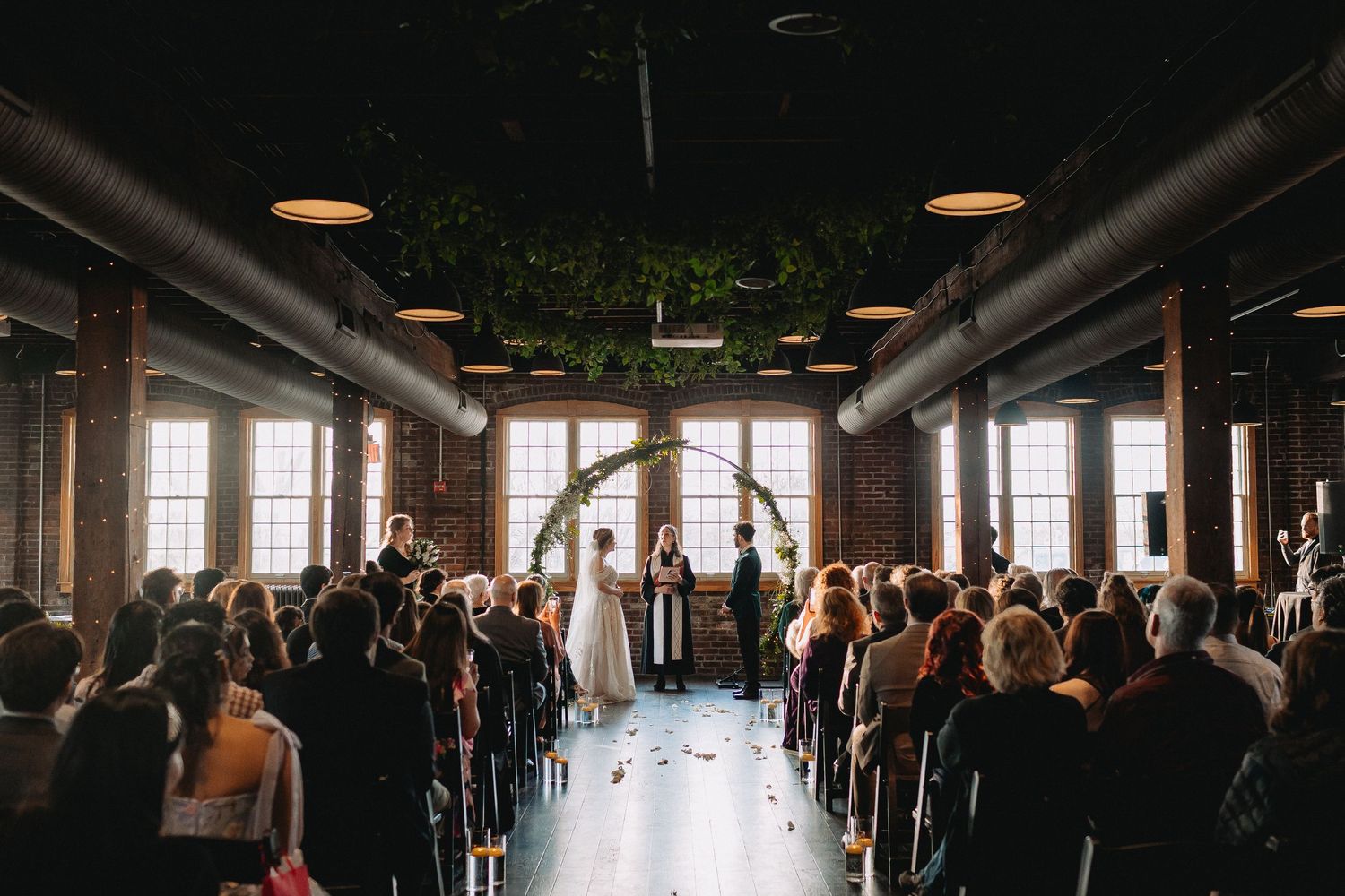 Wedding guests gather in a rustic industrial venue with exposed beams and large windows for an indoor celebration.