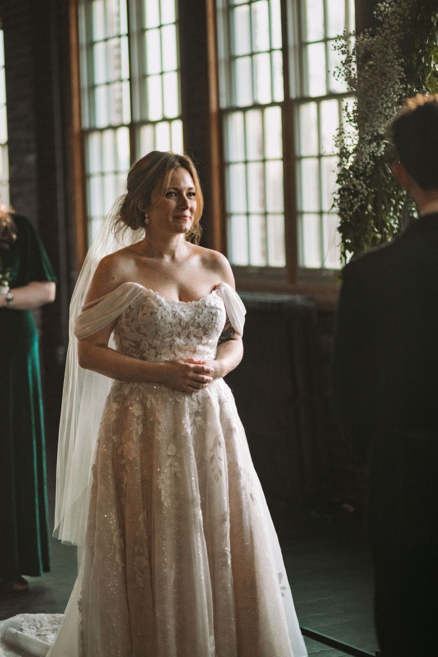A bride in a flowing lace dress stands in a sunlit room with tall windows.