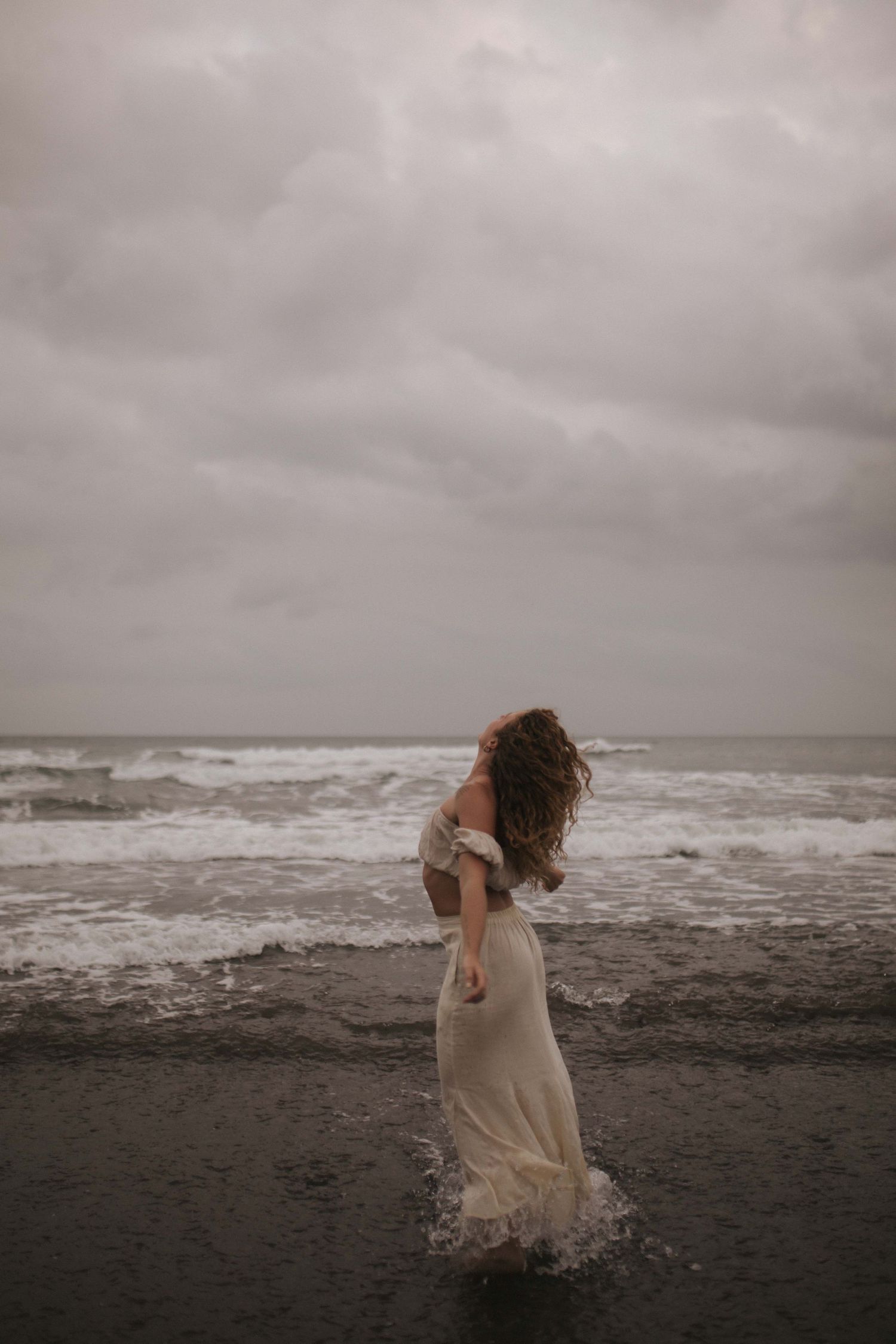 Woman in white dress running through ocean waves on dark beach under stormy clouds in New Zealand.