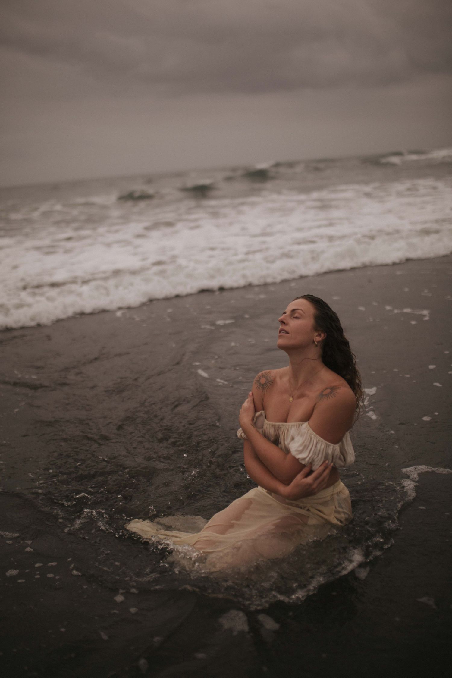 Woman in off-shoulder white dress kneeling in ocean surf with eyes closed in waves in New Zealand.