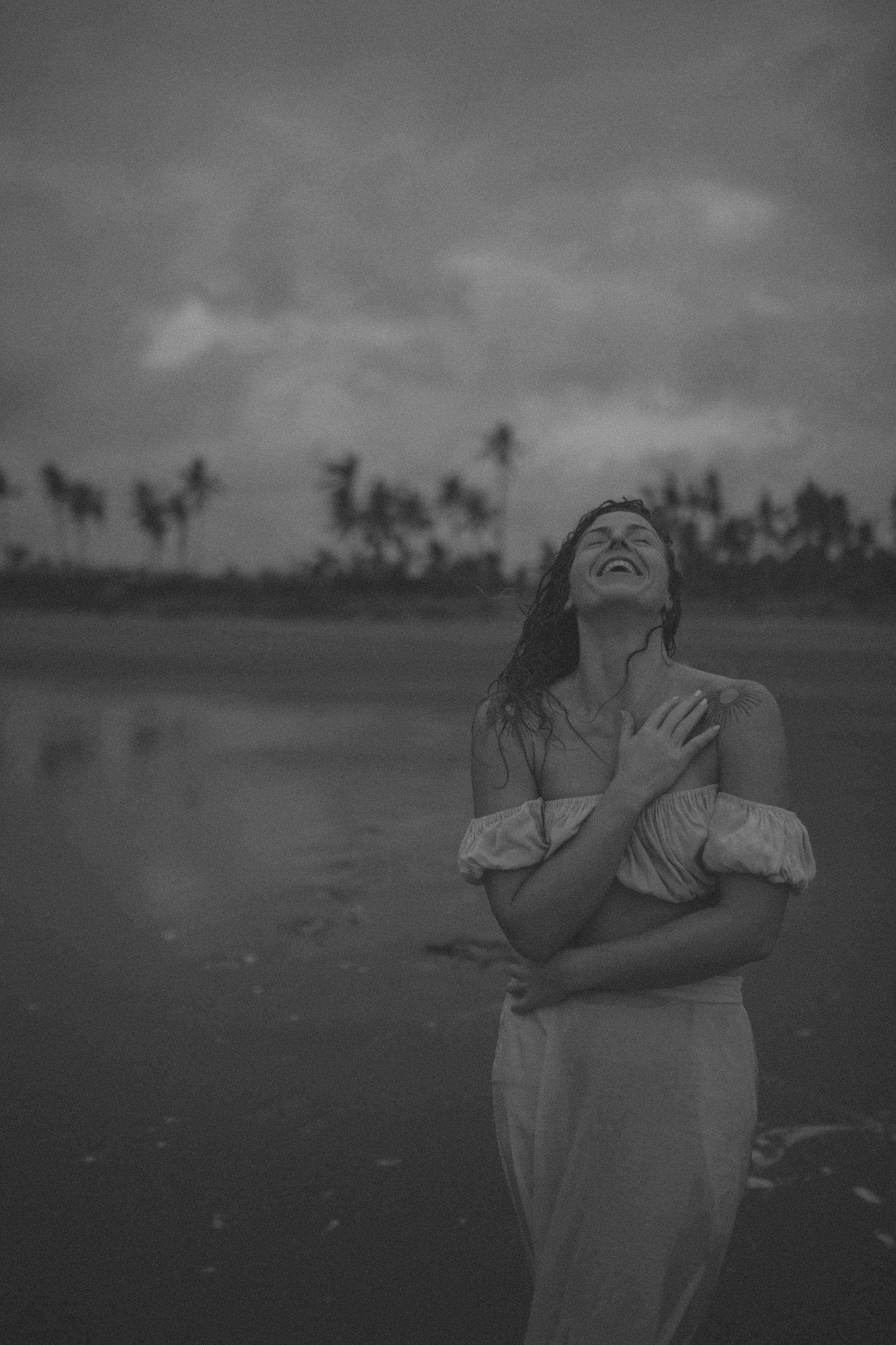 Black and white photo of a woman in a white dress laughing on a beach with palm trees in the background in New Zealand.