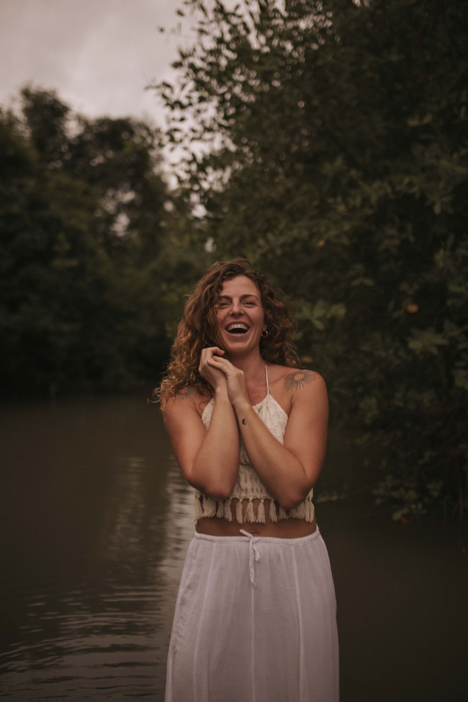 Joyful woman with curly hair laughing in white boho outfit standing near forest river in New Zealand.