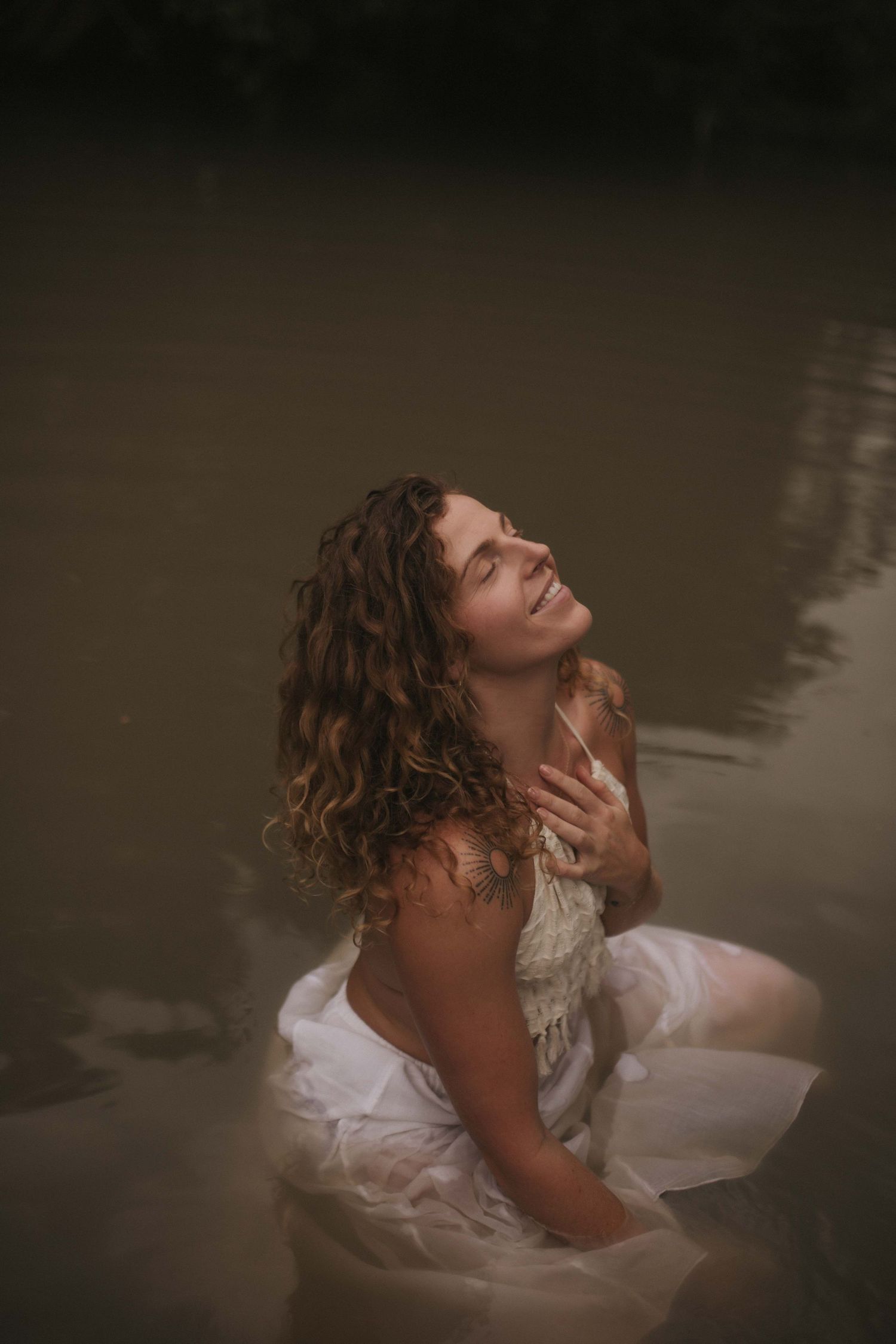 Woman in white dress sitting in calm river water with head tilted back peacefully in New Zealand.